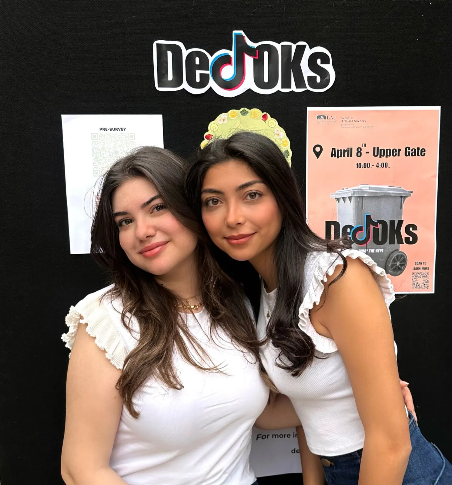 Joudi and Lea smiling in front of the DeTOKs booth wall, with the logo and the “April 8th · Upper Gate” poster.